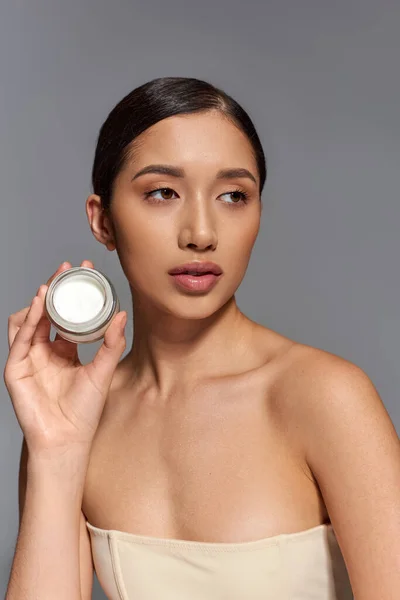 Product presentation, skin care, young asian model with brunette hair holding beauty jar with cream on grey background, glowing and heathy skin, beauty campaign, facial treatment concept — Stock Photo