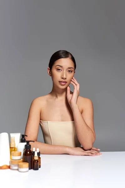 Produktpräsentation, Hautpflege, junges asiatisches Model mit brünetten Haaren posiert in der Nähe verschiedener Schönheitsprodukte auf grauem Hintergrund, glühende und heide Haut, Schönheitskampagne, Gesichtsbehandlungskonzept — Stockfoto