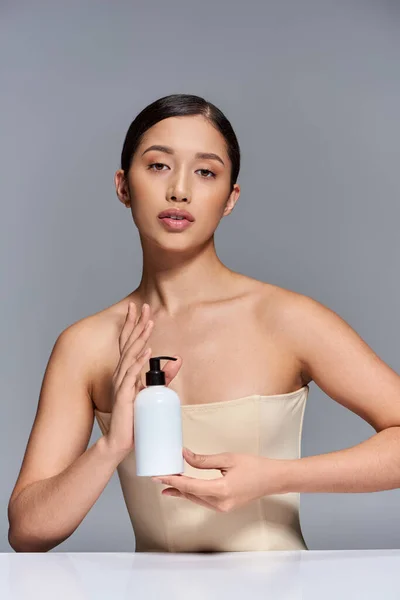 Beauty campaign, product presentation, young asian model with brunette hair holding cosmetic bottle with body lotion on grey background, glowing and heathy skin, facial treatment concept — Stock Photo