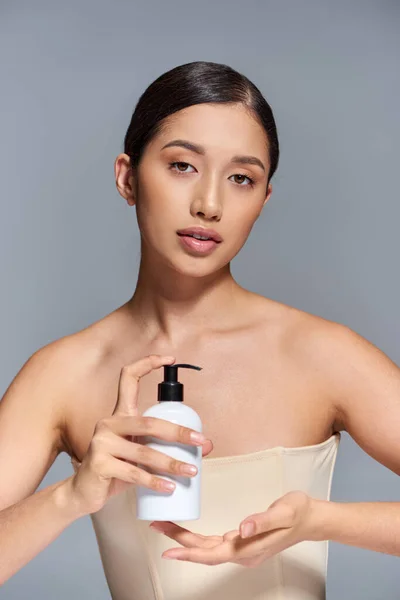 Косметическая кампания, презентация продукта, уход за кожей, молодая азиатская модель с брюнетками в руках косметическая бутылка на сером фоне, сияющая и вечнокожая кожа, концепция процедуры для лица — стоковое фото