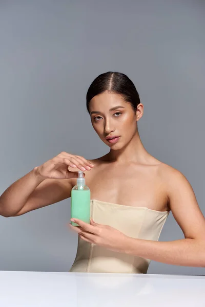 Produktpräsentation, Hautpflege, junges asiatisches Model mit brünetten Haaren mit Kosmetikflasche auf grauem Hintergrund, glühende und heide Haut, Schönheitskampagne, Gesichtsbehandlungskonzept — Stockfoto