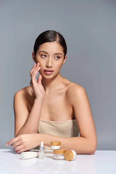Hautpflege-Präsentation, junge asiatische Frau mit nackten Schultern posiert in der Nähe verschiedener Schönheitsprodukte auf grauem Hintergrund, glühende und heide Haut, Schönheitskampagne, Gesichtsbehandlungskonzept — Stockfoto