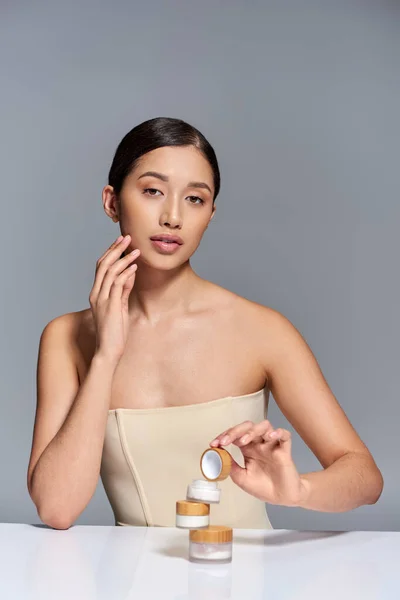 Produktpräsentation, Hautpflege, junges asiatisches Model mit brünetten Haaren, die Gesichtscreme auf grauem Hintergrund halten, glühende und heide Haut, Schönheitskampagne, Gesichtsbehandlung, konzeptionelle — Stockfoto