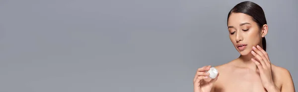 Cuidados com a pele, jovem mulher asiática com cabelo morena segurando frasco cosmético e aplicando creme de rosto no fundo cinza, brilhante e pagão pele, campanha de beleza, conceito de tratamento facial, banner — Fotografia de Stock