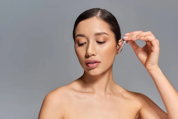Красива кампанія, догляд за шкірою, молода азіатська жінка з брюнеткою наносить сироватку з піпеткою на сірому фоні, світиться і погіршує шкіру, концепція лікування обличчя, дивлячись геть — стокове фото