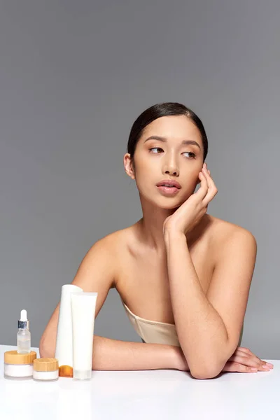 Schönheitsindustrie, junge asiatische Frau mit brünetten Haaren posiert in der Nähe von Schönheitsprodukten auf grauem Hintergrund, glühende und heide Haut, Gesichtsbehandlungskonzept, Gesichts- und Hautpflege — Stockfoto