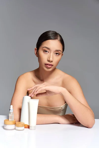 Schönheitsfotografie, junge asiatische Frau mit brünetten Haaren posiert in der Nähe von Schönheitsprodukten auf grauem Hintergrund, glühende und heide Haut, Gesichtsbehandlungskonzept, Gesichts- und Hautpflege, Jugend — Stockfoto