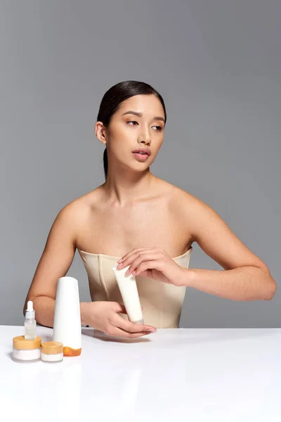 Кампания красоты, молодая азиатка с брюнетками, позирующая рядом с косметическими средствами на сером фоне, сияющая и вечнокожая кожа, концепция процедур для лица, уход за лицом и кожей, юность — стоковое фото