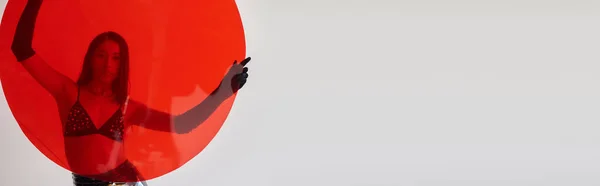 Fotografia di moda, stile lattice, giovane donna asiatica con capelli castani in posa in reggiseno e guanti e tenendo il vetro rotondo rosso su sfondo grigio, scelte di moda, dietro vetro, banner — Foto stock