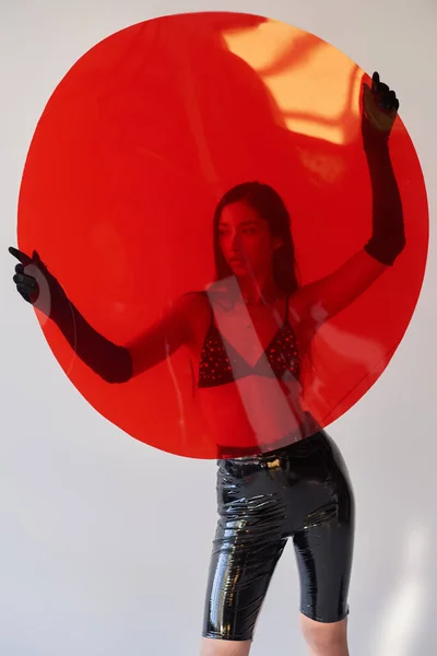 Opciones de moda, estilo látex, mujer asiática joven con cabello moreno posando en sujetador y guantes y sosteniendo vidrio rojo en forma redonda sobre fondo gris, opciones de moda, traje elegante, detrás de vidrio - foto de stock