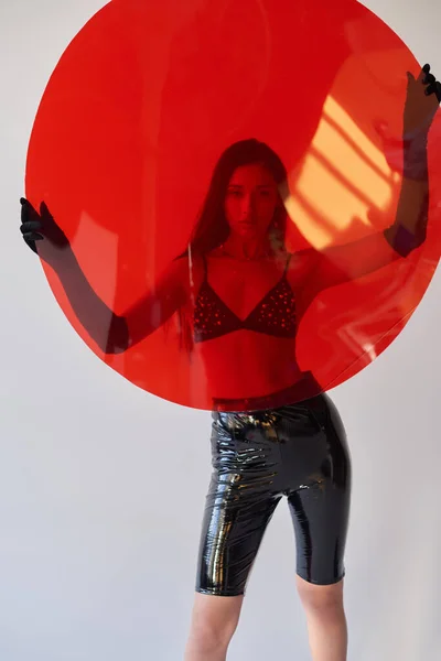 Fashion statement, latex style, young asian woman with brunette hair posing in bra and gloves while holding round shaped glass on grey background, fashion choices, stylish outfit, behind red glass — Stock Photo