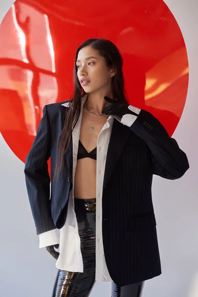 Edgy fashion, young asian woman in bra, white shirt and blazer posing in gloves near red round shaped glass, grey background, personal style, underwear and jacket, latex pants, youth — Stock Photo