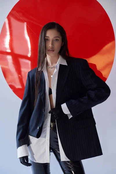 Moda vanguardista, mujer asiática joven en sujetador, camisa blanca y chaqueta posando en guantes cerca de vidrio redondo rojo, fondo gris, estilo personal, estilo látex, ropa interior y chaqueta, juventud - foto de stock