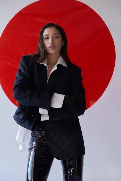 Модный выбор, молодая азиатская женщина в бюстгальтере, белая рубашка и блейзер позирует в перчатках возле красного круглого стекла, серый фон, личный стиль, нижнее белье и куртка, молодость — стоковое фото