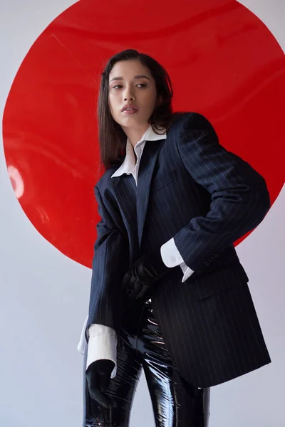 Mode avant, jeune femme asiatique en soutien-gorge, chemise blanche et blazer posant dans des gants près de verre rond rouge, fond gris, style personnel, pantalon en latex, sous-vêtements et veste, jeunesse — Photo de stock