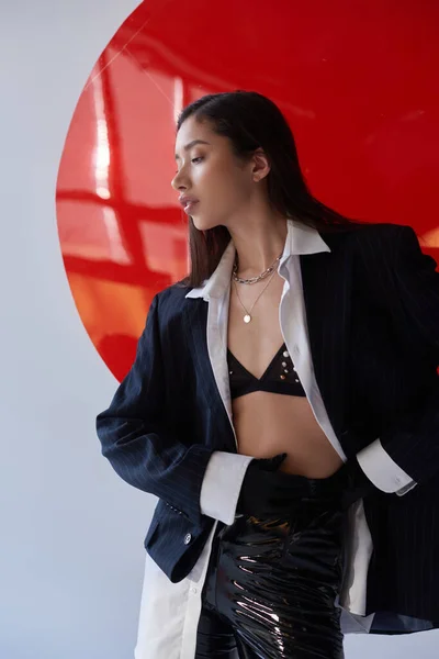 Moda vanguardista, mujer asiática joven en sujetador, camisa blanca y chaqueta posando en pantalones de látex y guantes negros y pantalones de látex cerca de vidrio redondo rojo, fondo gris, ropa interior y chaqueta - foto de stock