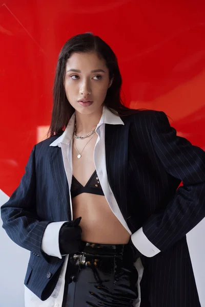 Стильный вид, молодая азиатская женщина в бюстгальтере, белая рубашка и блейзер позируют в перчатках возле красного круглого стекла, глядя в сторону на сером фоне, личный стиль, нижнее белье и куртка, молодость — стоковое фото