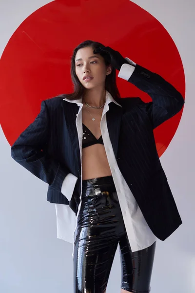 Stylish outfit, young asian woman in bra, white shirt and blazer posing in gloves and latex shorts near red round shaped glass, looking away on grey background, personal style, youth — Stock Photo