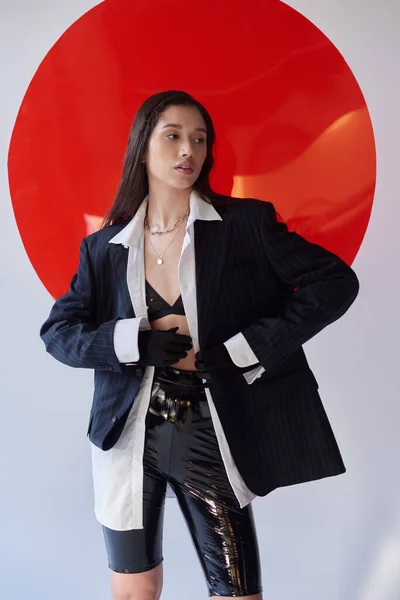 Fashion forward, young asian model in bra, white shirt and blazer posing in gloves and latex shorts near red round shaped glass, grey background, looking away, personal style, youth trend — Stock Photo