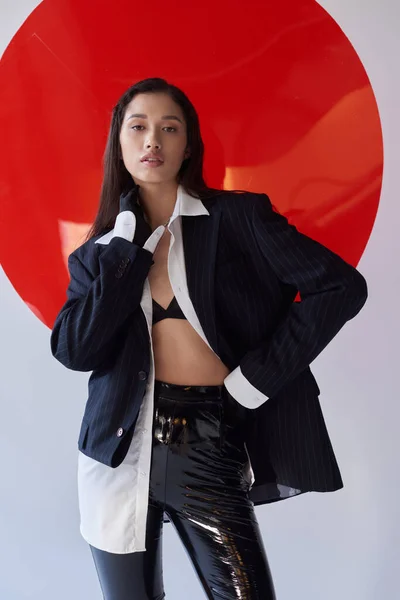 Fashion photography, young asian model in bra, white shirt and blazer posing in gloves and latex shorts near red round shaped glass, grey background, looking away, personal style, youth trend — Stock Photo
