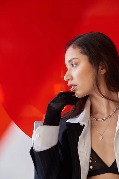 Moderno individuo, fotografía de moda, joven modelo asiático en camisa blanca y chaqueta posando en guantes cerca de vidrio redondo rojo, fondo gris, mirando hacia otro lado, estilo personal, tendencia - foto de stock