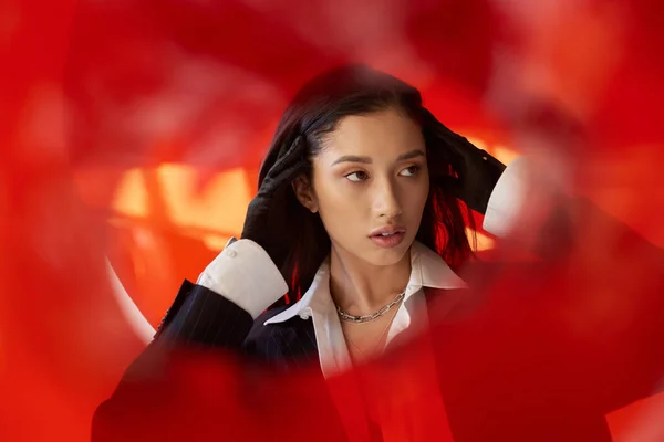 Moda adelante, joven modelo asiático en camisa blanca y chaqueta posando en guantes detrás de cristal rojo, estilo personal, tendencia juvenil, conceptual, cuello conmovedor y posando en estudio - foto de stock