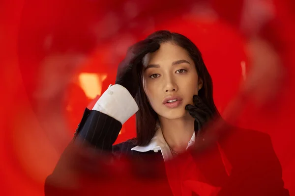 Estilo personal, fotografía de moda, joven modelo asiático en camisa blanca y chaqueta posando en guantes cerca de vidrio redondo rojo, fondo gris, mirando a la cámara, estilo moderno, tendencia juvenil - foto de stock