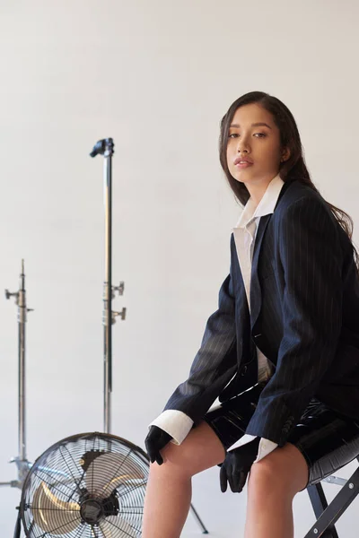 Studiofotografie, junge asiatische Frau im Blazer, weißem Hemd und Latex-Shorts sitzt auf Klappstuhl neben elektrischem Ventilator auf grauem Hintergrund, Mode und Stil, blickt in die Kamera — Stockfoto