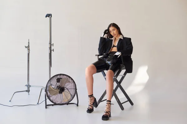Studiofotografie, junge asiatische Frau im Blazer, weißem Hemd und Latex-Shorts auf Klappstuhl neben elektrischem Ventilator auf grauem Hintergrund sitzend, modisches Statement, wegschauen, in voller Länge — Stockfoto