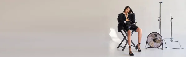 Studio fotografia, giovane donna asiatica in blazer, camicia bianca e pantaloncini di lattice seduto sulla sedia pieghevole vicino ventilatore elettrico su sfondo grigio, dichiarazione di moda, guardando altrove, lunghezza intera, banner — Foto stock