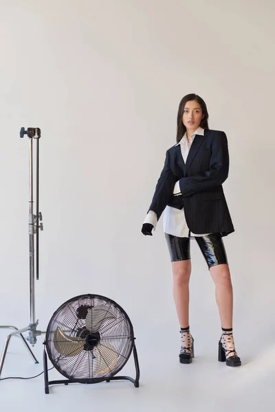 Moda para a frente, fotografia de estúdio, jovem mulher asiática em olhar elegante posando em fundo cinza, blazer, camisa branca e calções de látex, pé perto de ventilador elétrico, estilo pessoal, comprimento total — Fotografia de Stock