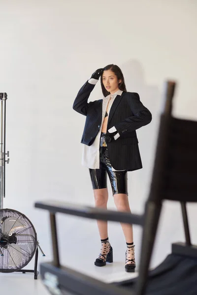 Opciones de moda, fotografía de estudio, mujer asiática joven en aspecto elegante posando sobre fondo gris, chaqueta, camisa blanca y pantalones cortos de látex, de pie cerca de ventilador eléctrico, estilo personal, longitud completa - foto de stock