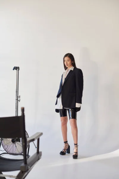 Edgy fashion, studio photography, junge asiatische Frau in stylischem Look posiert auf grauem Hintergrund, Blazer, weißes Hemd und Latex-Shorts, neben elektrischem Ventilator stehend, persönlicher Stil, volle Länge — Stockfoto