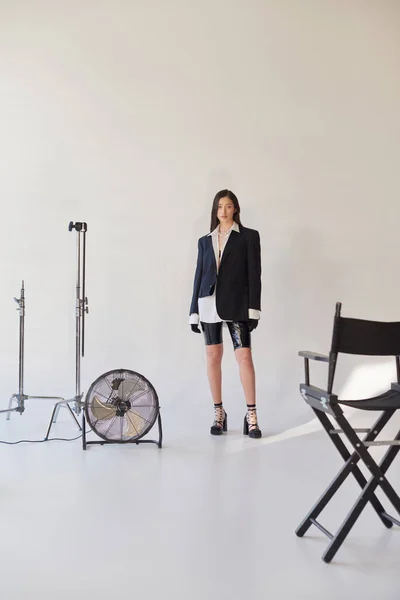 Mode und Stil, Studiofotografie, junge asiatische Frau in stylischem Look posiert auf grauem Hintergrund, Blazer, weißes Hemd und Latex-Shorts, neben elektrischem Ventilator stehend, persönlicher Stil, volle Länge — Stockfoto