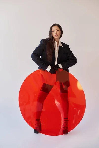 Stile spigoloso, fotografia di studio, giovane donna asiatica in look elegante posa vicino a vetro rotondo rosso, sfondo grigio, blazer e pantaloncini di lattice, moda giovanile, lunghezza intera — Foto stock