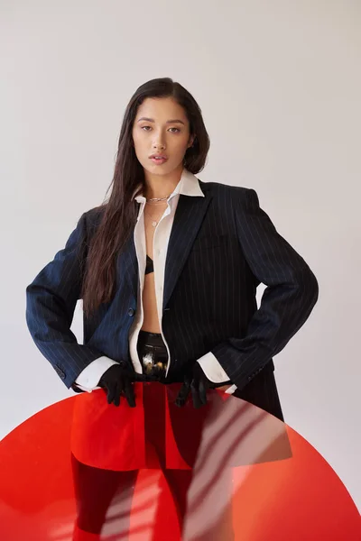 Estilo vanguardista, fotografía de estudio, mujer asiática joven en aspecto elegante posando cerca de vidrio redondo rojo, fondo gris, blazer y pantalones cortos de látex, moda juvenil, estilo fresco - foto de stock
