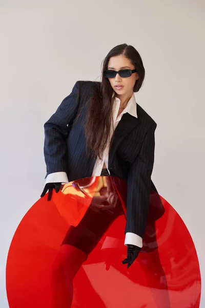 Estilo vanguardista, fotografía de estudio, mujer asiática joven en aspecto elegante y gafas de sol posando cerca de vidrio redondo rojo, fondo gris, blazer y pantalones cortos de látex, moda juvenil, estilo fresco - foto de stock