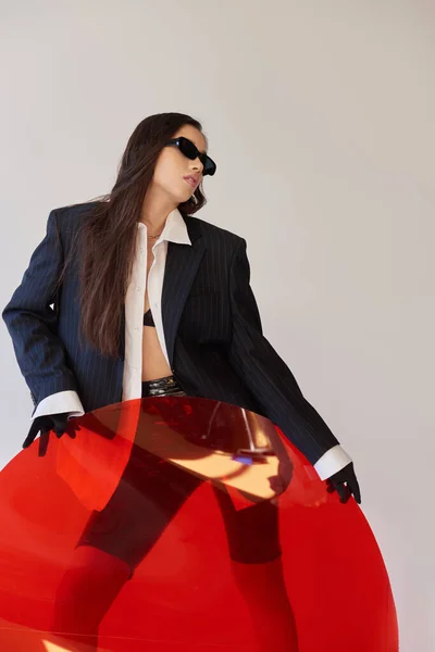 Moda moderna, fotografía de estudio, mujer asiática joven en aspecto elegante y gafas de sol posando cerca de vidrio redondo rojo, fondo gris, blazer y pantalones cortos de látex, moda juvenil, estilo fresco - foto de stock