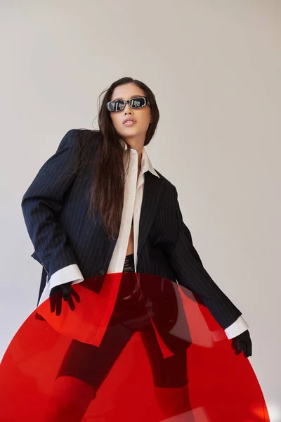 Резкий стиль, студийная фотография, молодая азиатская модель в стильном виде и солнцезащитные очки, позирующие возле красного круглого стекла, серый фон, пиджак и латексные шорты, молодая мода, современная женщина — стоковое фото
