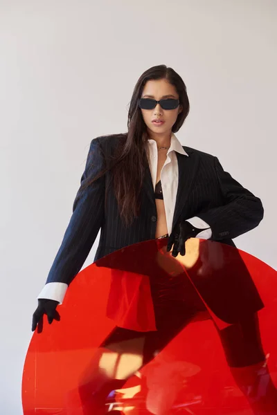 Adelante de la moda, fotografía de estudio, modelo asiático joven en aspecto elegante y gafas de sol posando cerca de vidrio redondo rojo, fondo gris, blazer y pantalones cortos de látex, moda juvenil, mujer moderna - foto de stock