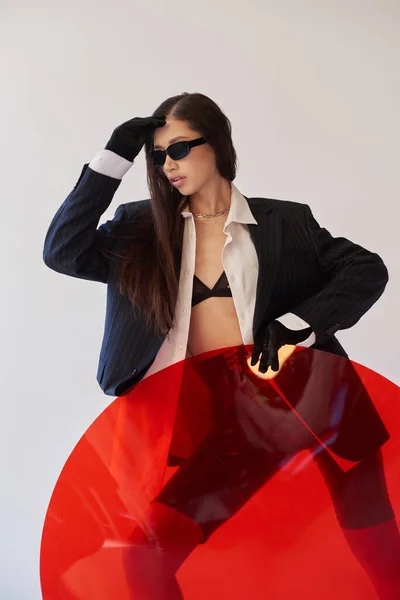 Cooler Stil, Studiofotografie, junges asiatisches Model im stylischen Look und Sonnenbrille, die in der Nähe von rotem Rundglas posiert, grauer Hintergrund, Blazer und Latex-Shorts, jugendliche Mode, moderne Frau — Stockfoto