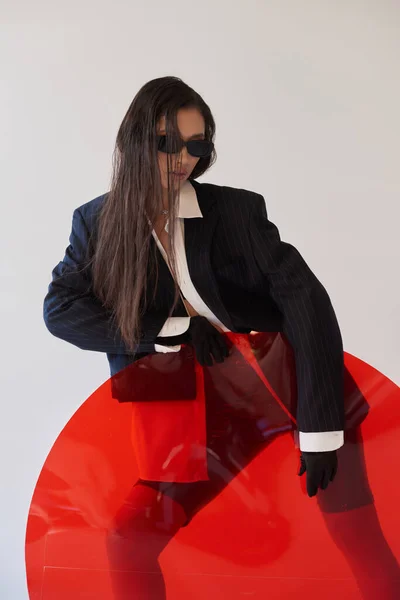 Moda de látex, modelo asiático joven en aspecto elegante y gafas de sol posando cerca de vidrio redondo rojo, fondo gris, blazer y pantalones cortos de látex, moda juvenil, mujer moderna, fotografía de estudio - foto de stock