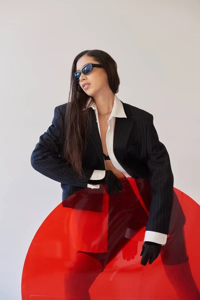 Schöne asiatische Modell in stilvollem Look und Sonnenbrille posiert in der Nähe von rotem Rundglas, grauen Hintergrund, Blazer und Latex-Shorts, jugendliche Mode, moderne Frau, kantigen Stil, Studiofotografie — Stockfoto