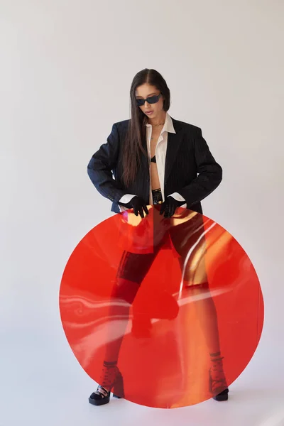 Attractive asian model in stylish look and sunglasses posing holding red round shaped glass, grey background, blazer and latex shorts, youthful and modern woman, fashion forward, studio photography — Stock Photo