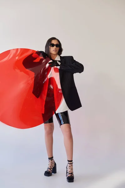 Hübsches asiatisches Model in stylischem Look und Sonnenbrille posiert mit rotem Rundglas, grauem Hintergrund, Blazer und Latex-Shorts, jugendliche und moderne Frau, modisches Statement, Studiofotografie — Stockfoto
