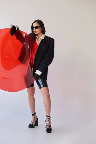 Красивая азиатская женщина в стильном виде и солнцезащитные очки в позе держа красное круглое стекло, серый фон, блейзер и латексные шорты, молодая модель, мода вперед, студийная фотография — стоковое фото