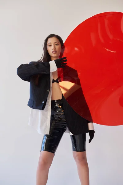 Красивая азиатская женщина в модном наряде, держа красное круглое стекло, серый фон, пиджак и черные латексные шорты, молодая модель в перчатках, мода вперед, студийная фотография, концептуальный — стоковое фото