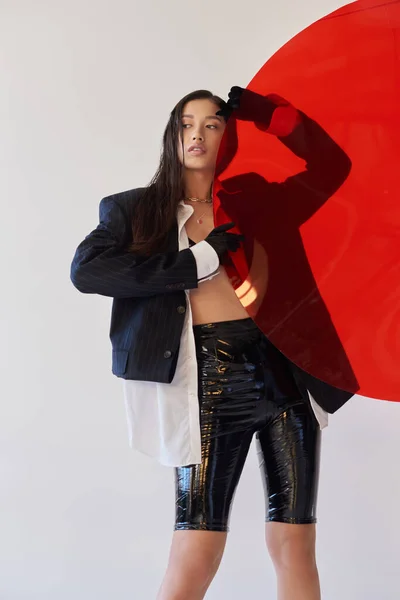 Bonita mujer asiática en traje de moda sosteniendo vidrio redondo rojo, fondo gris, blazer y pantalones cortos de látex negro, modelo joven en guantes, la moda hacia adelante, fotografía de estudio, conceptual - foto de stock