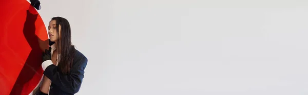 Jolie femme asiatique en tenue tendance tenant verre rond rouge, fond gris, blazer et short en latex noir, modèle jeune en gants, choix de mode, photographie studio, conceptuel, bannière — Photo de stock