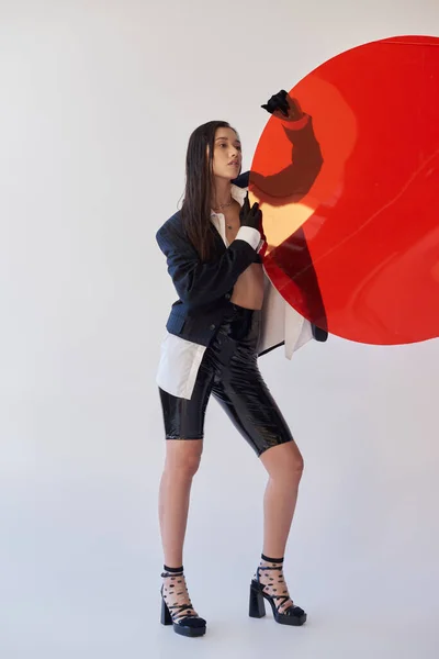 Bonita mujer asiática en traje de moda sosteniendo vidrio redondo rojo, fondo gris, blazer y pantalones cortos de látex negro, modelo juvenil en guantes, moda y estilo, fotografía de estudio, conceptual - foto de stock